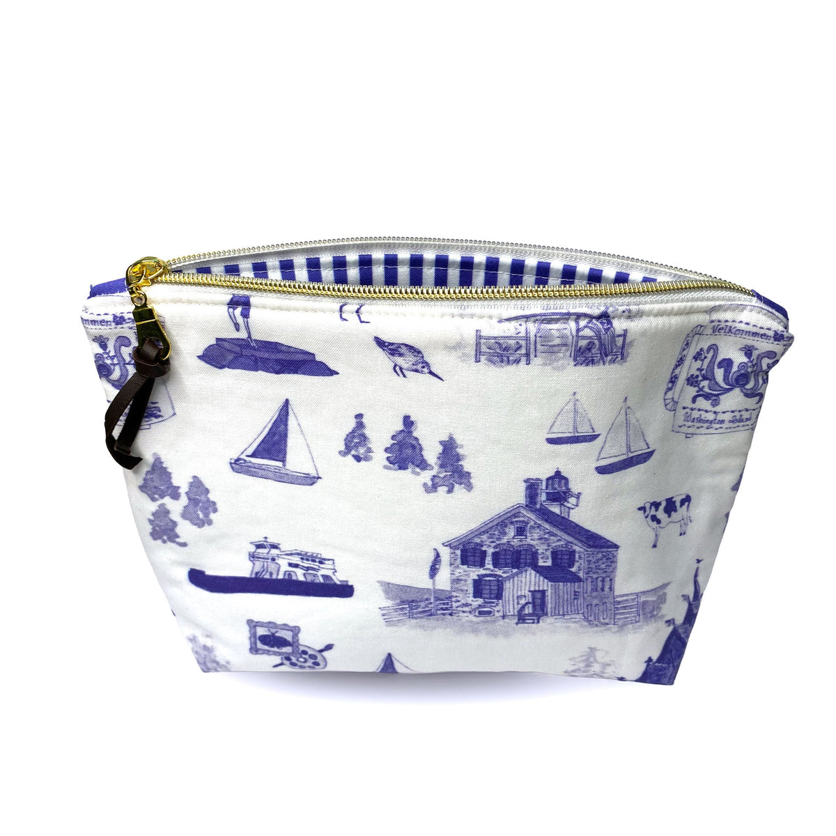 A zippered pouch with a white fabric body and a blue washington island toile pattern featuring sailboats, a lighthouse, and a cow, ferry, coffee pot. It has a gold zipper, a blue and white striped interior lining, and a brown leather zipper pull.